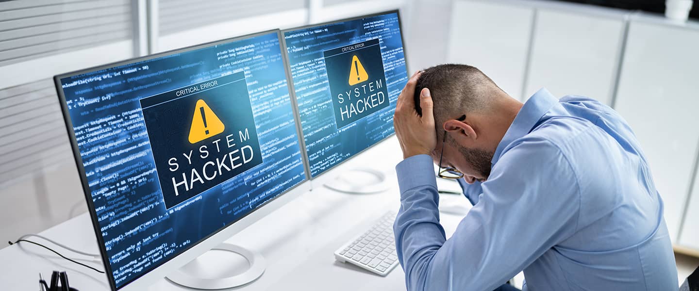 A man sitting in front of his computer with his head in his hands and a system hacked message appearing on the screen.