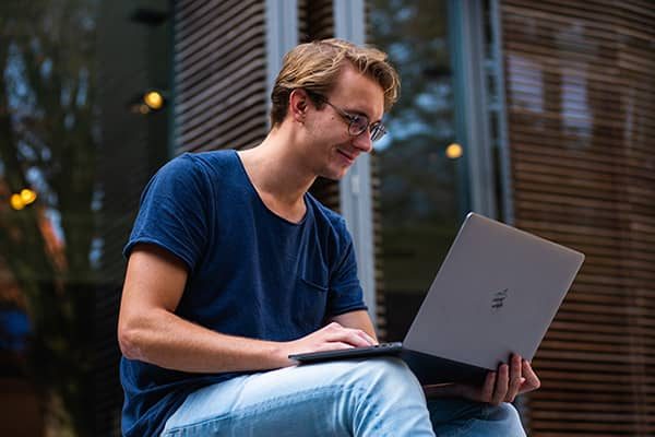 person wearing glasses, blue tshirt and jeans sitting outside while working on his laptop